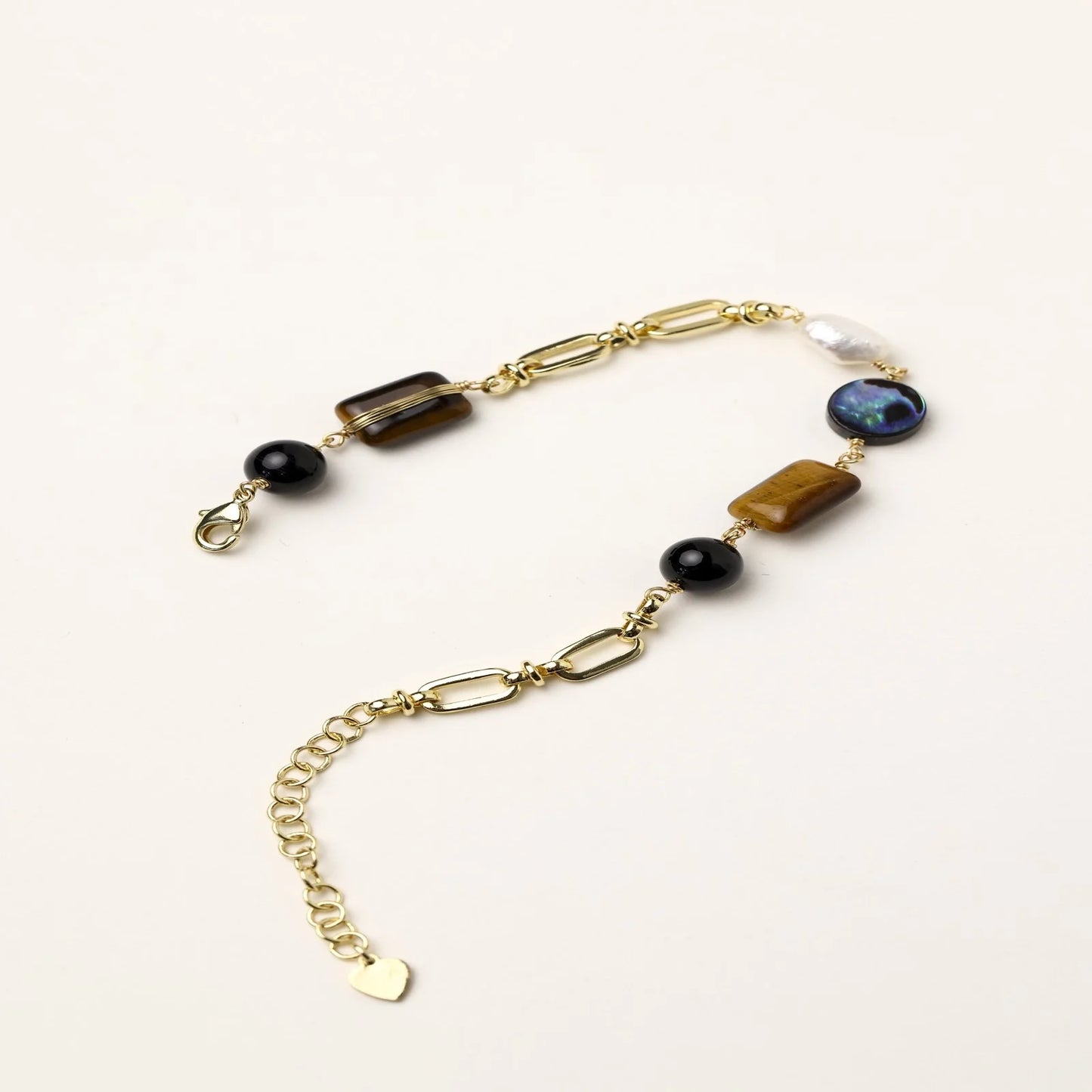Gold chain bracelet with tiger's eye, obsidian, pearl, and iridescent abalone stones on cream background.