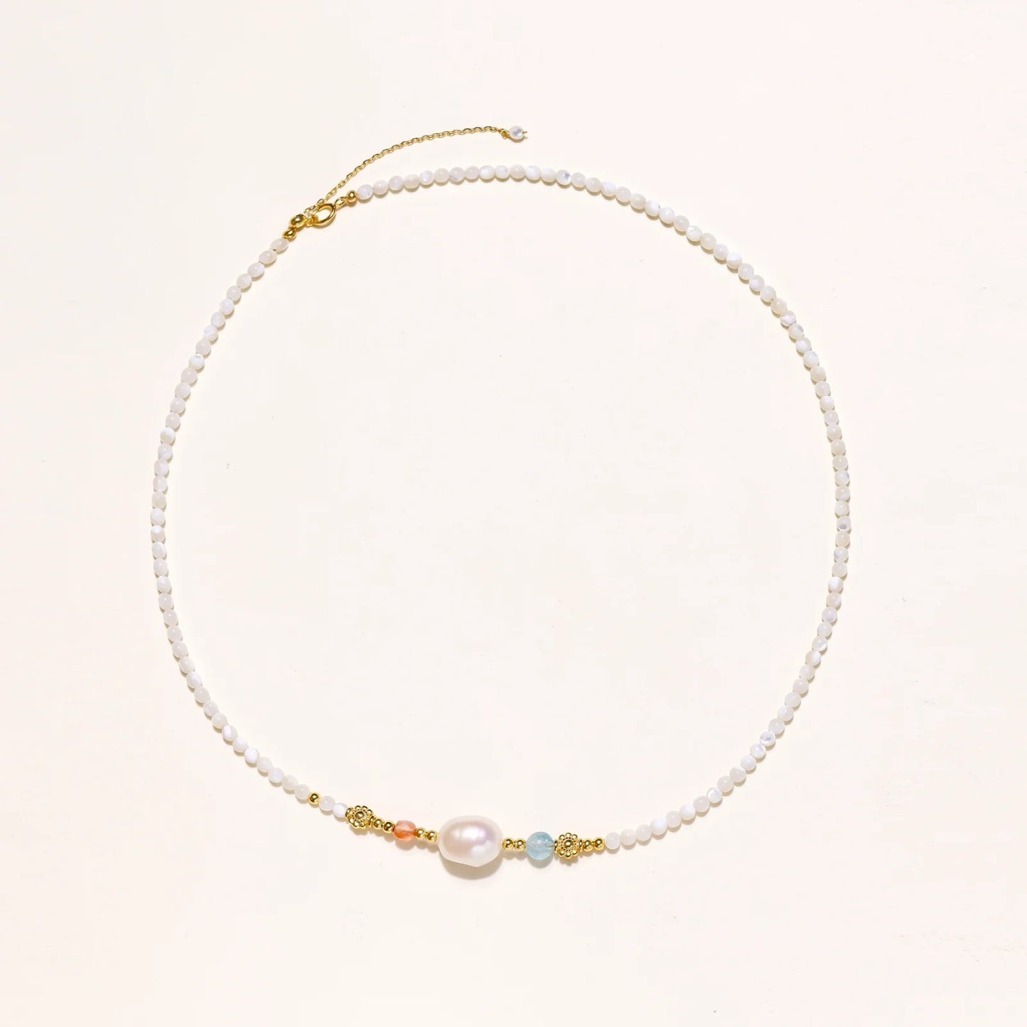 Delicate white beaded necklace with baroque pearl center, gold accents, and coral and aqua gemstones on cream background.