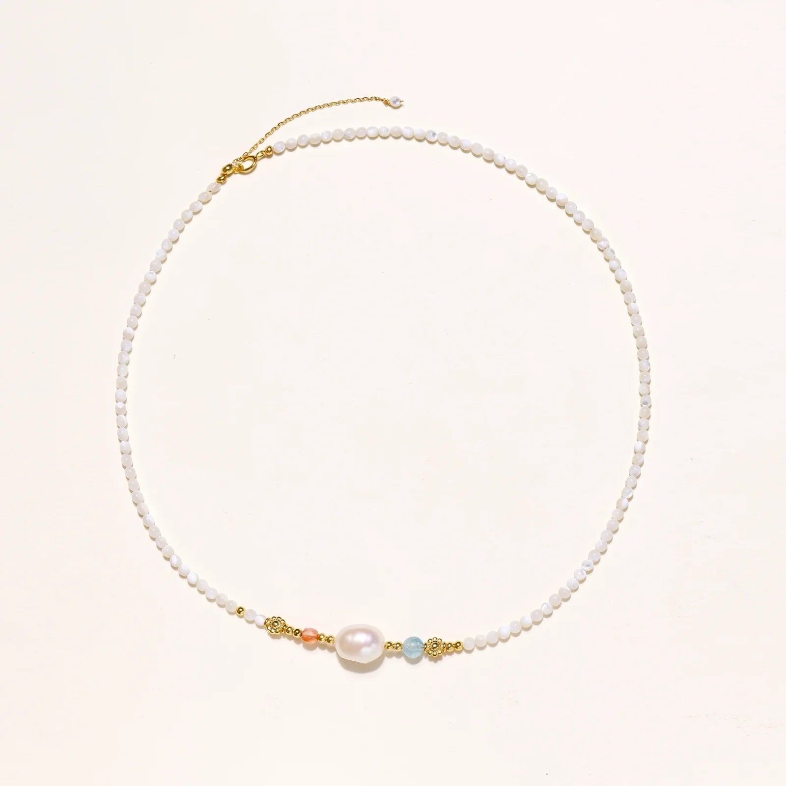 Delicate white beaded necklace with baroque pearl center, gold accents, and coral and aqua gemstones on cream background.