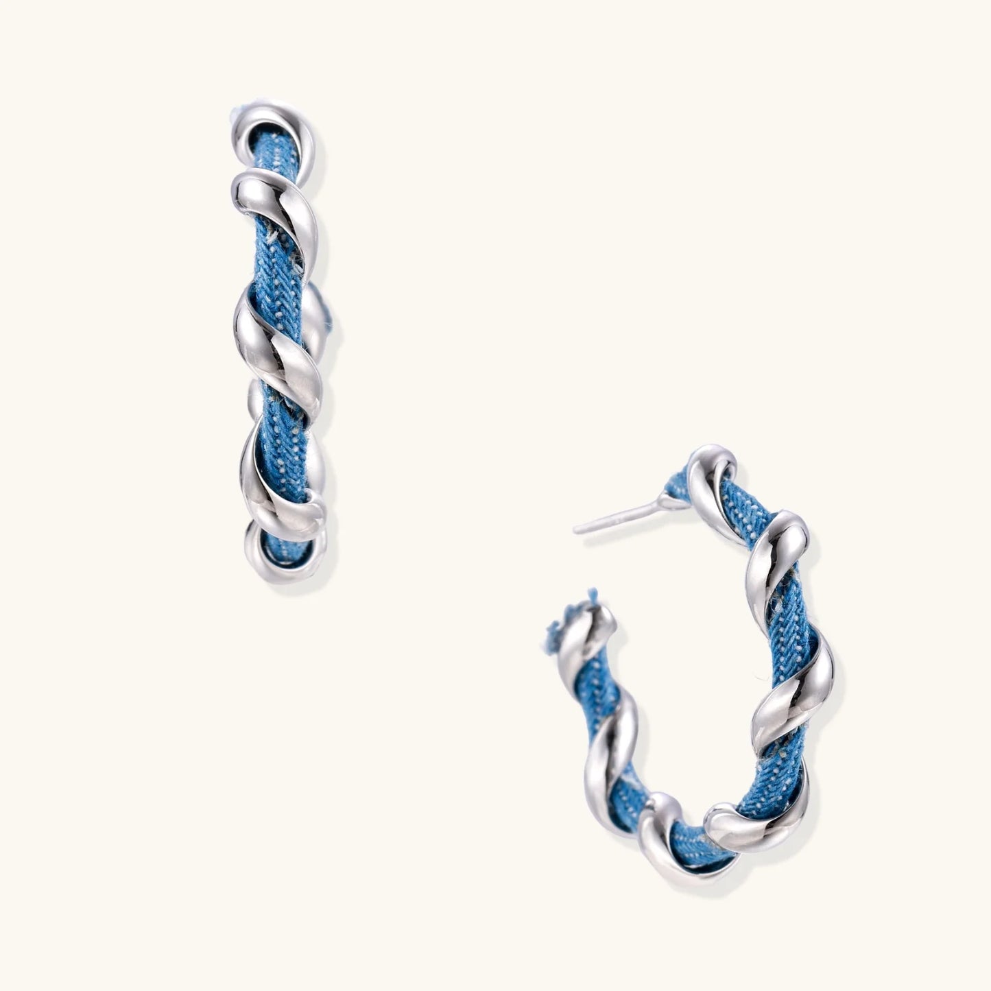 White gold-plated hoops intertwined with blue denim fabric in a twisted rope design on cream background.