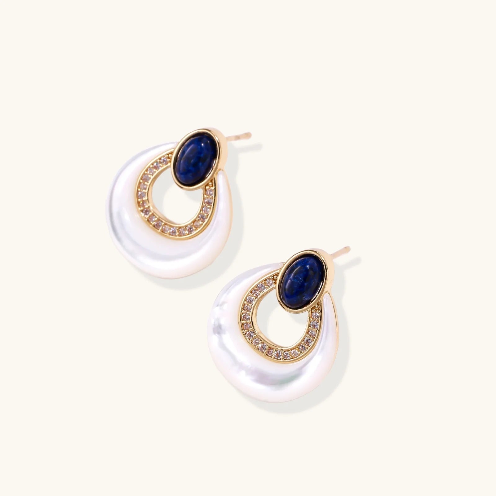 Gold drop earrings with oval lapis lazuli, crystal-paved frame, and iridescent mother-of-pearl teardrop on cream background