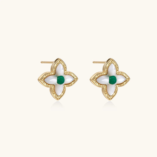 Gold quatrefoil stud earrings with white mother-of-pearl petals and emerald green gemstone center on beige background.