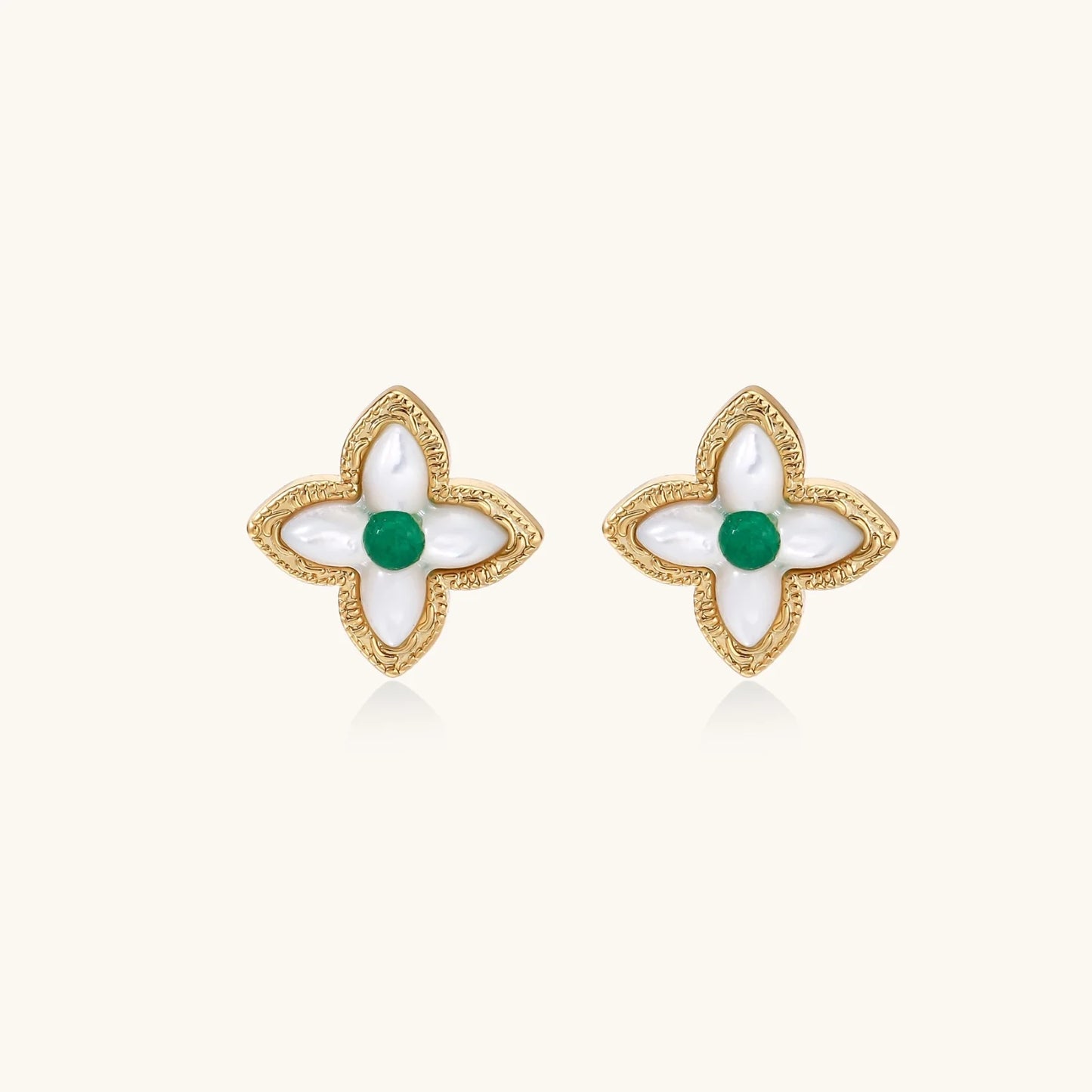 Gold quatrefoil stud earrings with white mother-of-pearl petals and emerald green gemstone center on beige background.