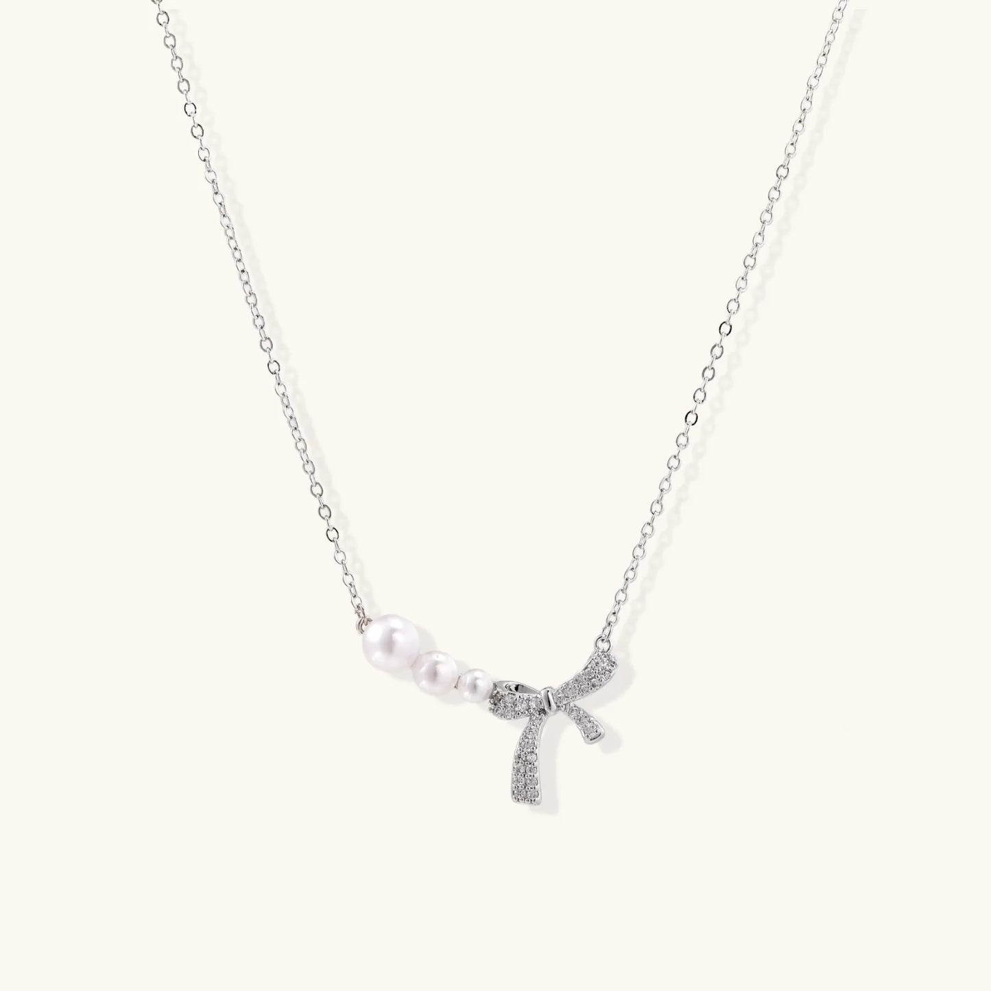 Delicate white gold plated necklace with graduated freshwater pearls and crystal-studded bow pendant on ivory background