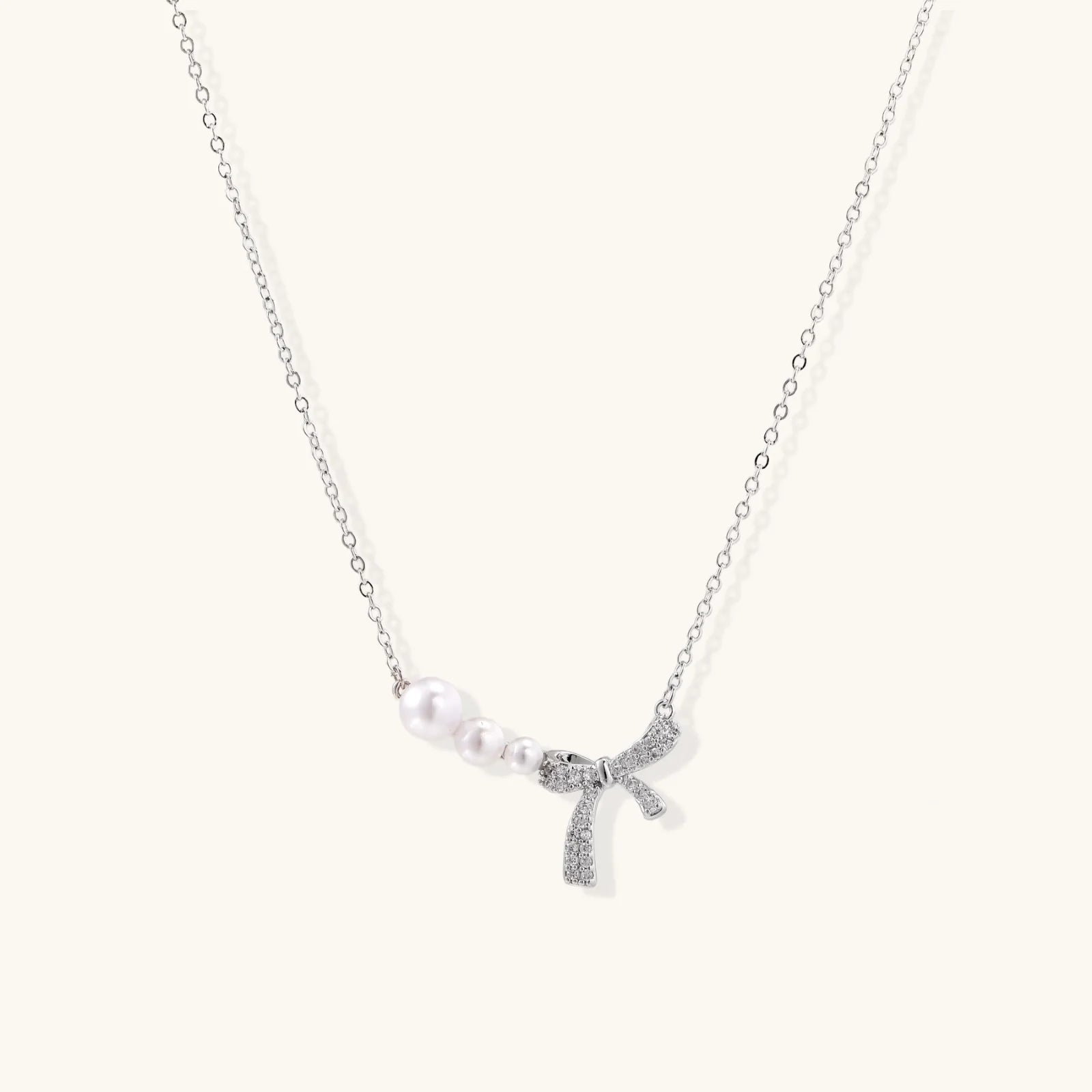 Delicate white gold plated necklace with graduated freshwater pearls and crystal-studded bow pendant on ivory background