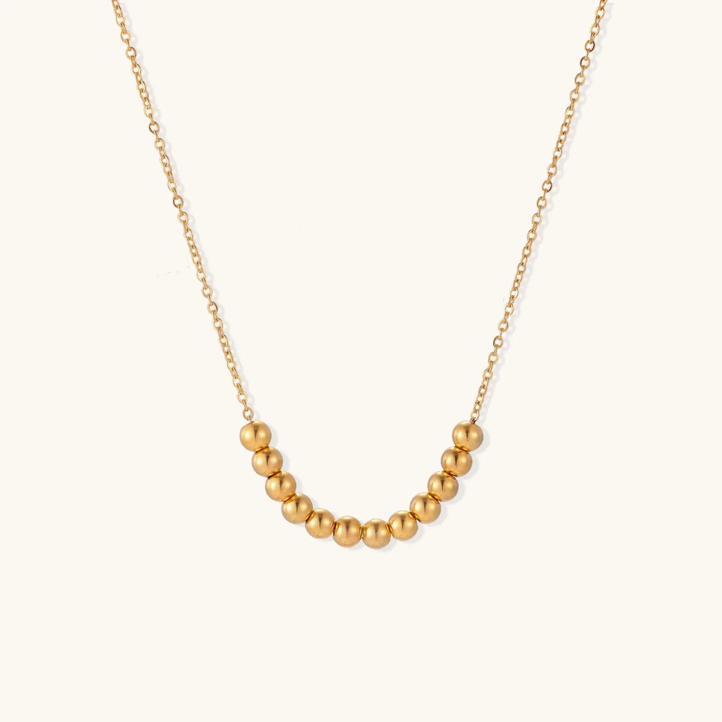 Delicate gold-plated necklace with graduated spherical beads forming gentle smile curve on beige background